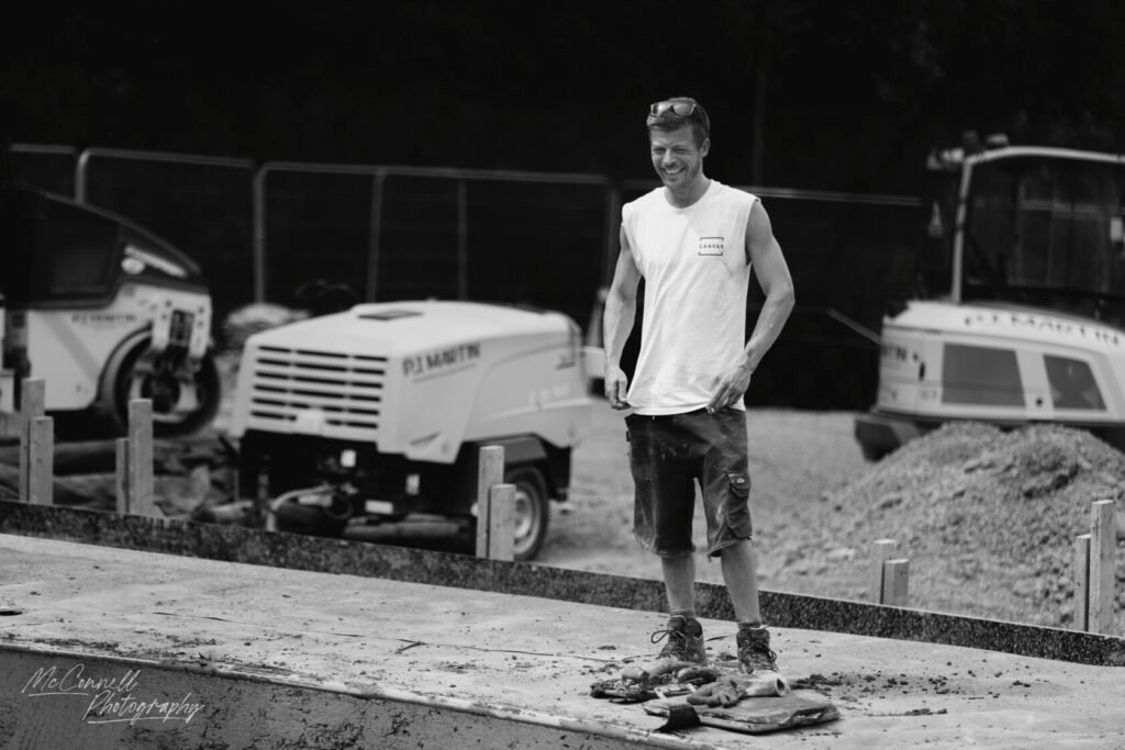 A young male construction worker standing on a concrete surface, smiling, wearing a sleeveless white shirt and work trousers, with construction machinery in the background.