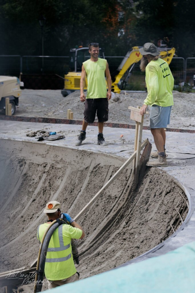Three construction workers in bright yellow vests pouring concrete into a curved surface with machinery in the background.