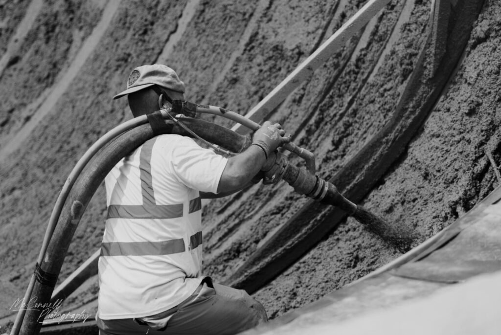 A construction worker in a reflective vest using a concrete sprayer to apply material on a surface.