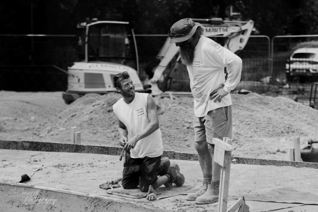 Two construction workers, one kneeling and smiling while holding tools, and another standing with a beard and hat, on a building site with machinery in the background.