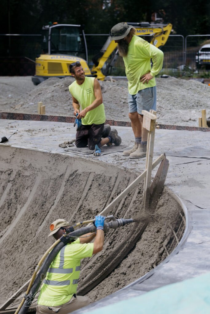Three construction workers in bright yellow shirts are working on a project, with one kneeling and pouring concrete into a curved form while the others observe.