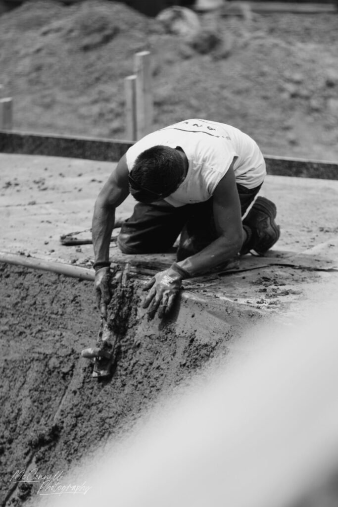 A construction worker kneeling on the ground, using a trowel to smooth wet concrete in a construction site.
