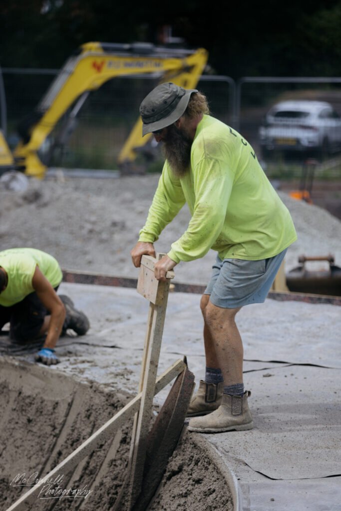 A construction worker wearing a bright yellow shirt and hat, using a wooden tool to shape wet concrete on a construction site, with another worker in the background.