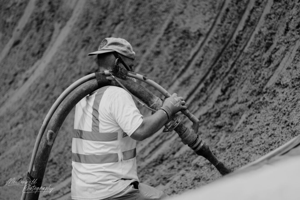 A construction worker in a reflective vest is handling a hose, with sand in the background, captured in black and white.