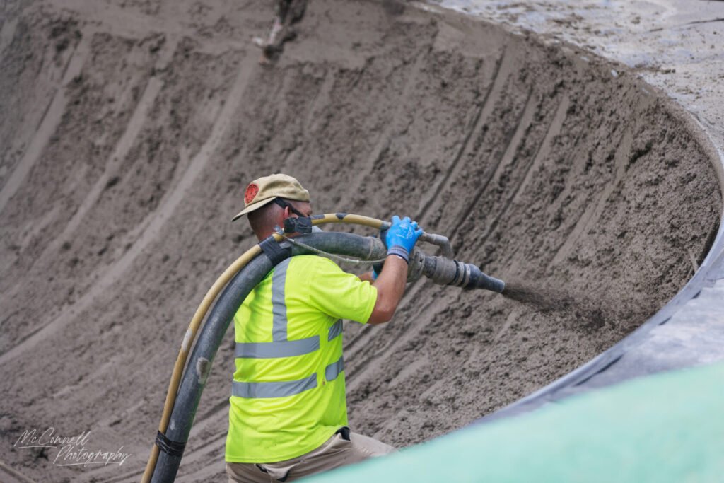 A construction worker wearing a bright yellow safety vest and gloves operates a concrete pump to pour concrete into a curved surface.