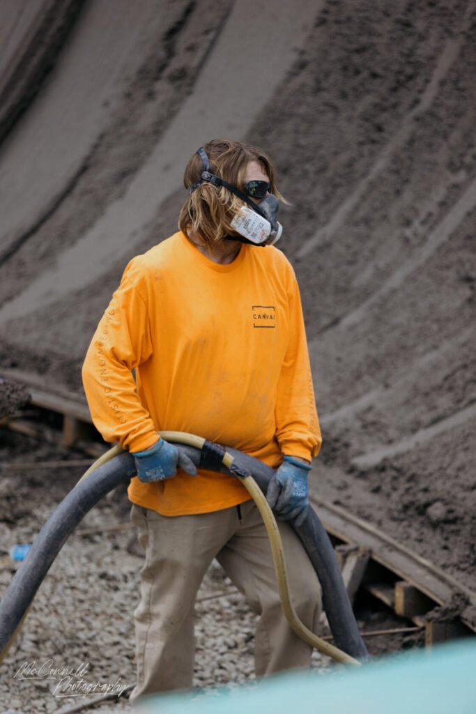 A construction worker wearing a bright orange long-sleeve shirt and a respirator holds a flexible hose at a worksite covered in grey material.