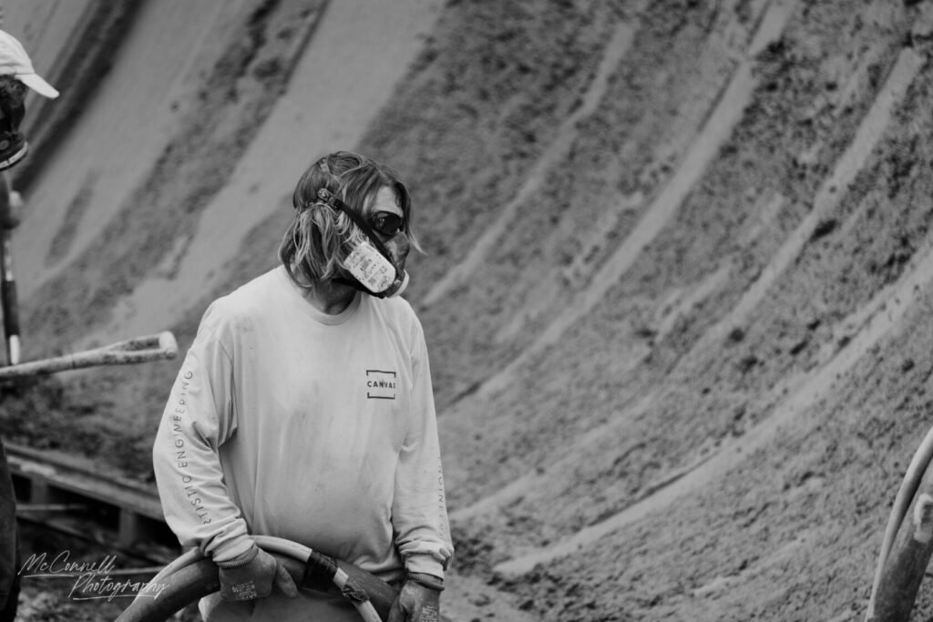 A construction worker with long hair wearing sunglasses and a mask, holding a hose while standing on a construction site with sandy textures in the background.