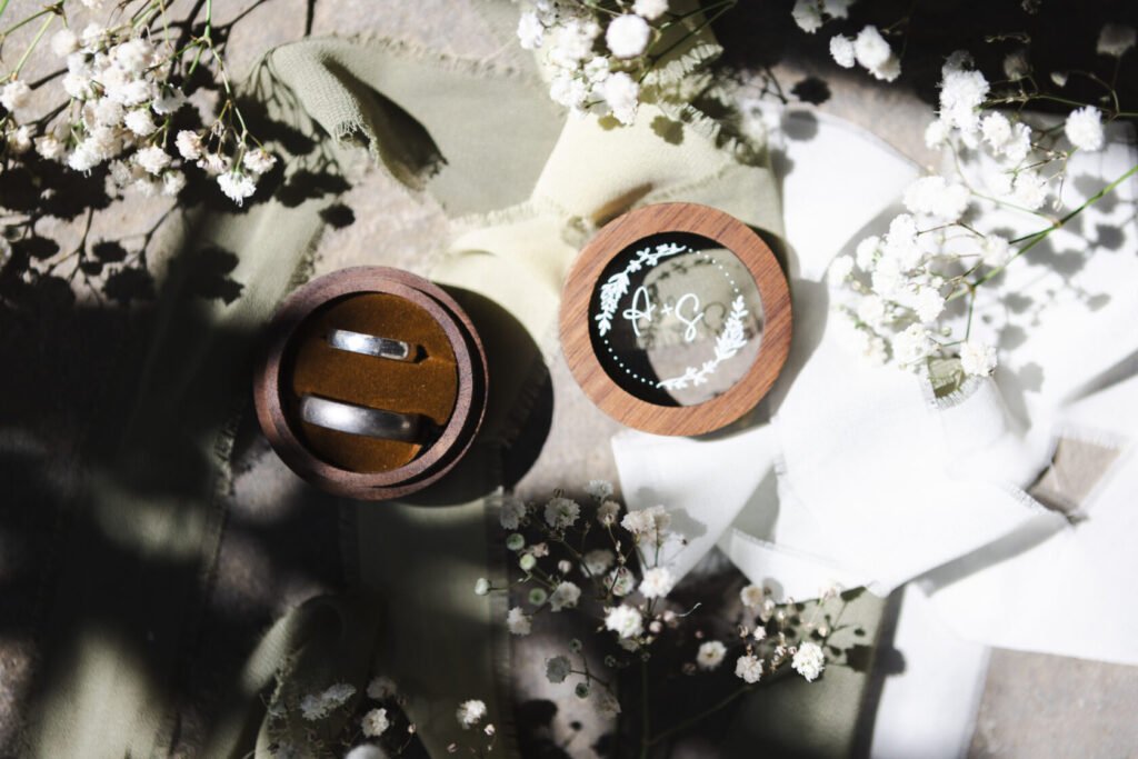 Two silver wedding rings displayed in a round wooden box, surrounded by delicate white flowers and soft fabric in muted green and cream tones.