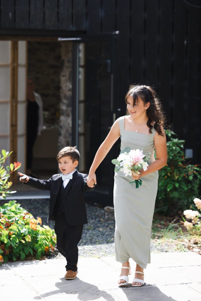 A young boy in a black suit holds hands with a woman in a light green dress, both walking together. The boy is smiling and pointing, while the woman carries a bouquet of flowers. They are in a garden setting with a dark wooden building in the background.