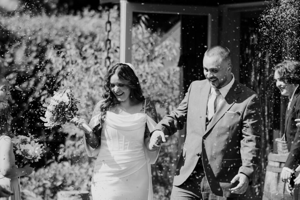 A happy couple, newlyweds, walking hand in hand while confetti is falling around them. The bride wears a white dress and holds a bouquet, while the groom is in a grey suit. Friends and family are celebrating in the background.