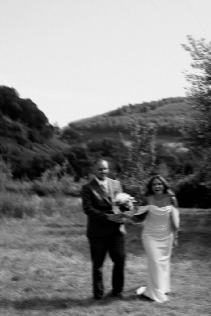 A blurred photograph of a man and woman joyfully walking hand in hand through a grassy field, with mountains in the background. The woman is wearing a white dress and holds a bouquet.
