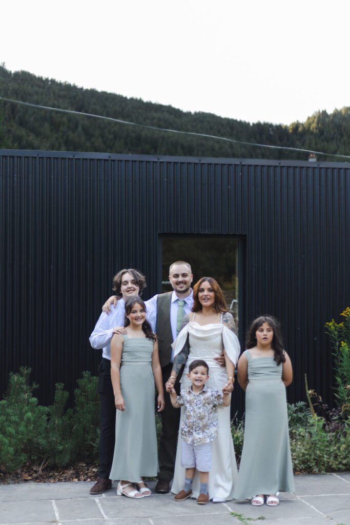 A family poses outdoors in front of a modern black building. The group includes five children and two adults, with the adults dressed in formal attire and children in matching green dresses and a floral shirt.