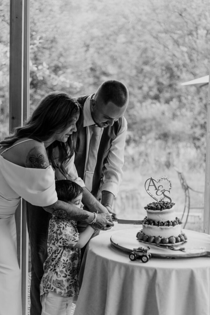 A woman, man, and child are joyfully cutting a wedding cake together. The cake is decorated with strawberries and a personalised cake topper featuring 'A&S'.
