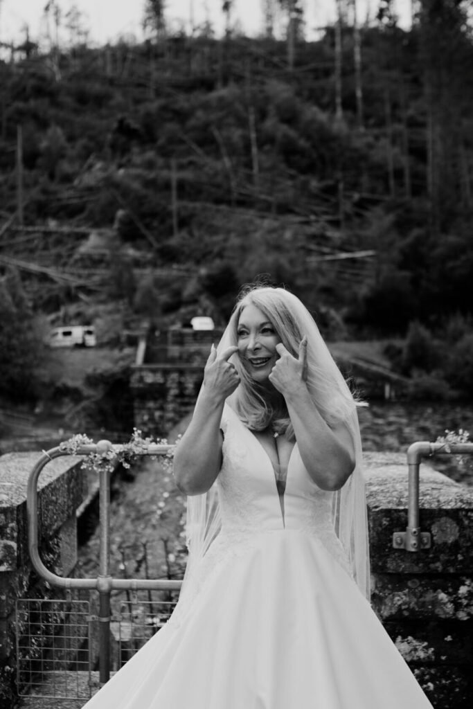 A bride with long blonde hair, wearing a white wedding dress, playfully touching her face with both hands, smiling against a backdrop of trees.