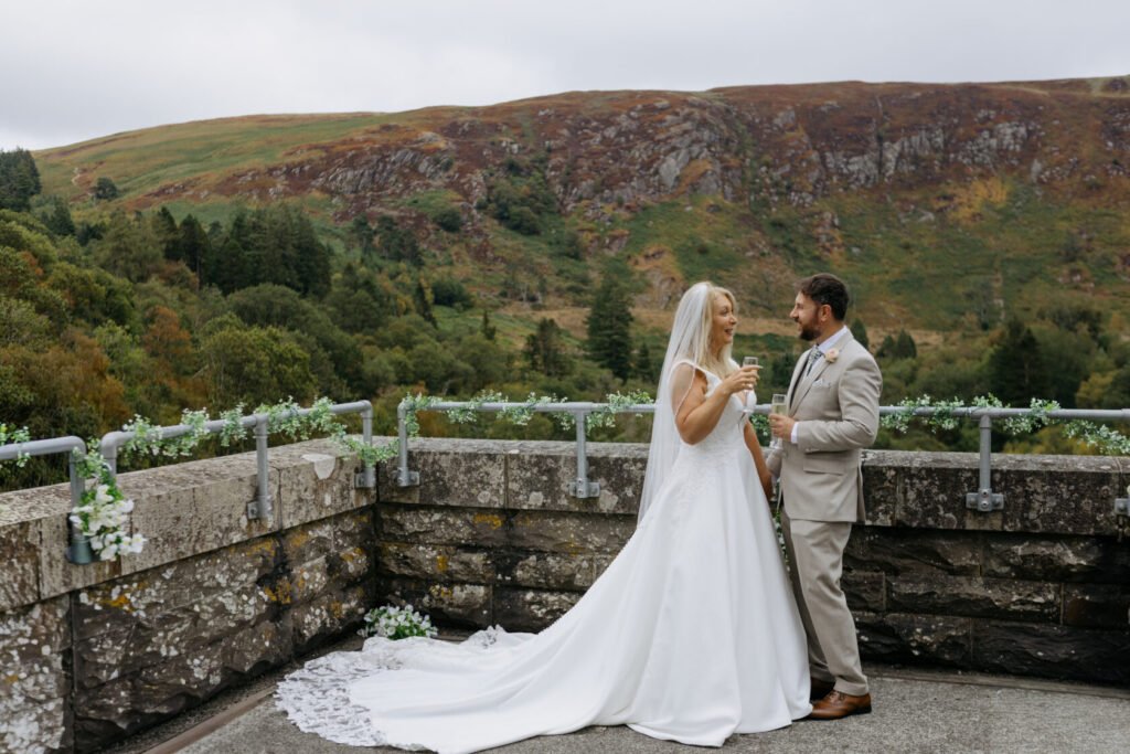A happy couple celebrating their wedding, standing on a stone terrace with a scenic mountain landscape in the background. The bride wears a flowing white gown and veil, while the groom is dressed in a light suit. Both hold glasses of champagne.