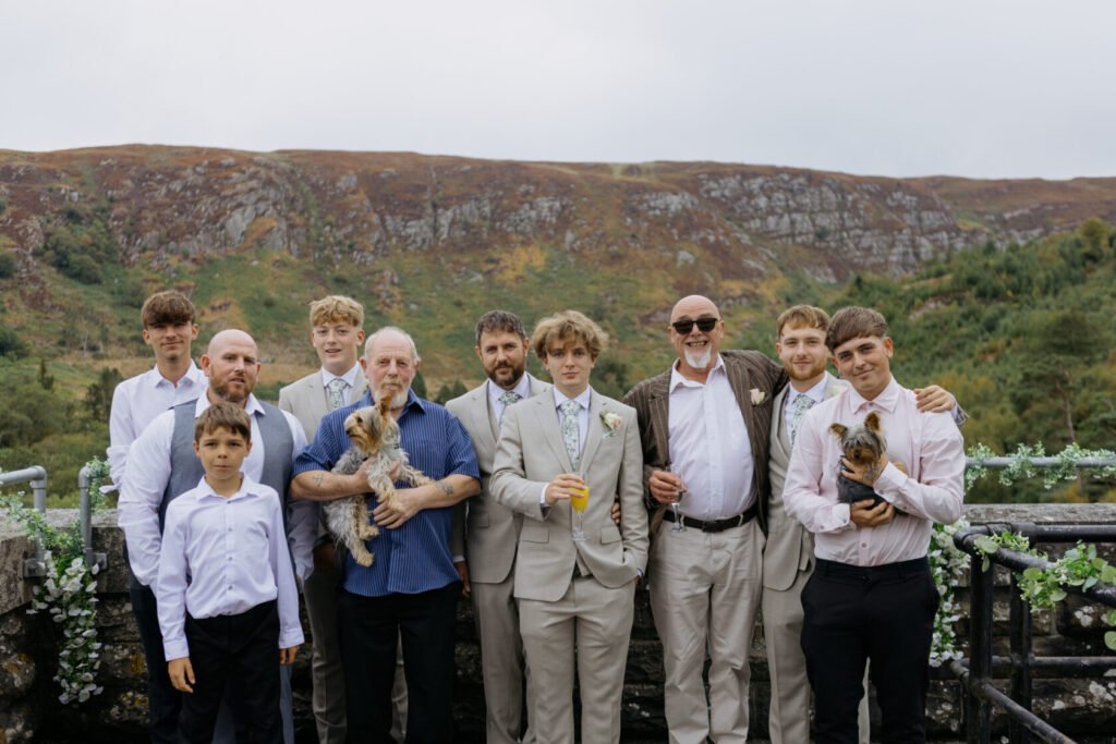 A group of men and boys, dressed in formal attire, pose together for a wedding photo, with two small dogs in their arms and a scenic backdrop of hills.
