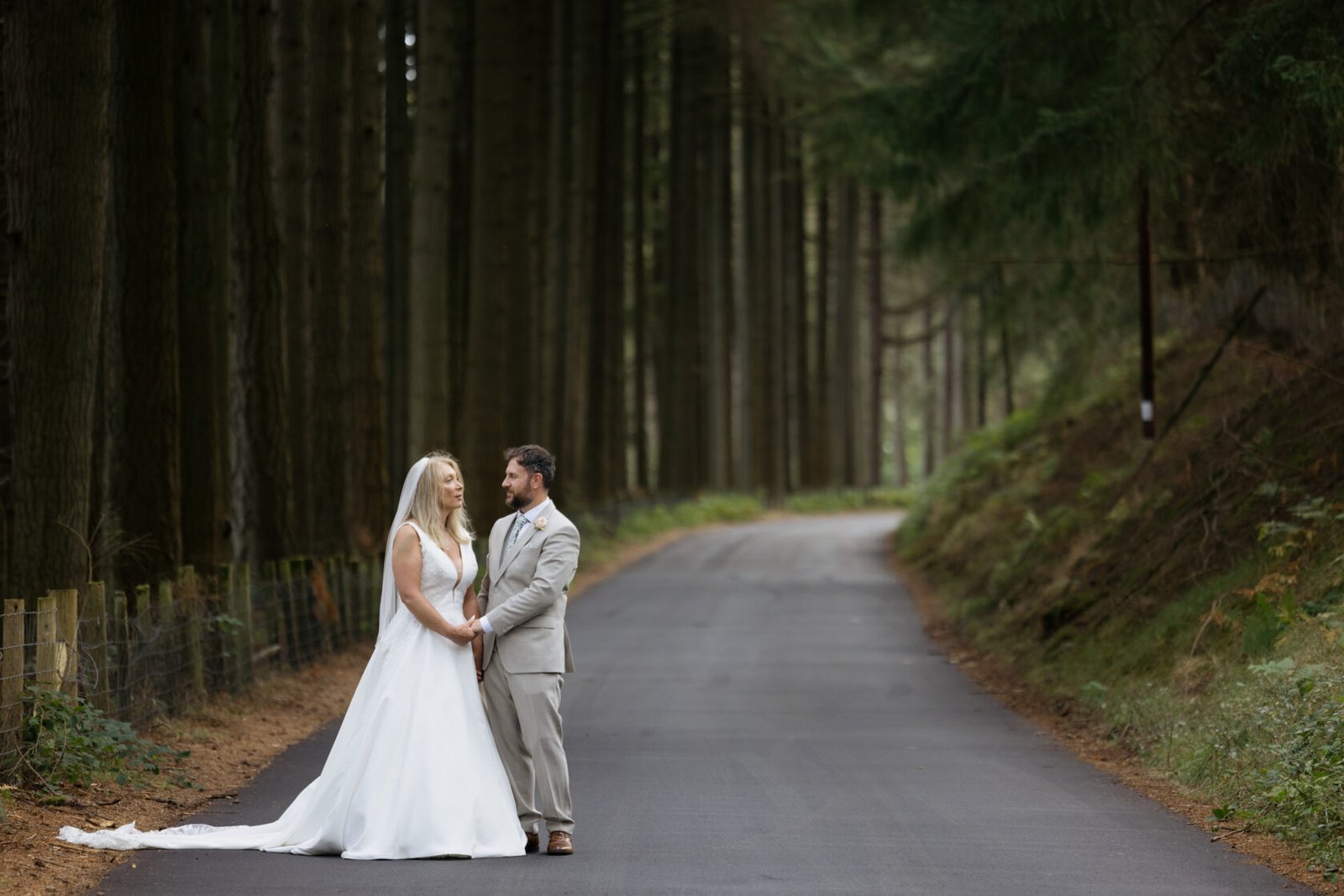 A bride in a white gown and a groom in a light suit holding hands on a tranquil forest road, surrounded by tall trees.