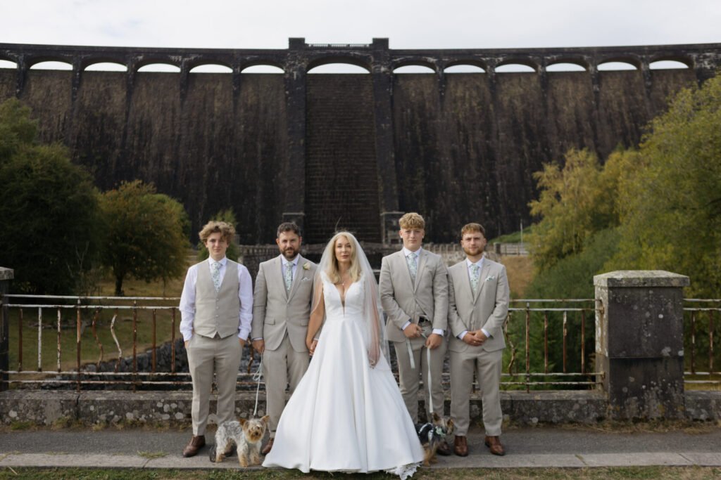 A bride in a white gown stands with four men dressed in light grey suits, two of whom hold leashes for small dogs, in front of a large dam with a stone wall.