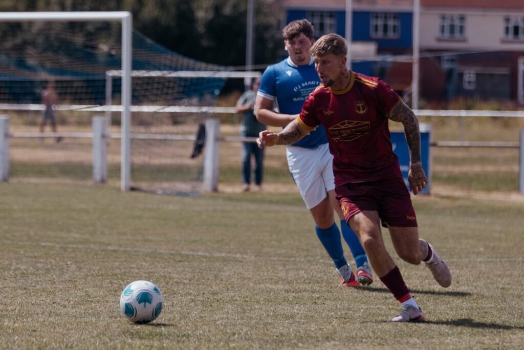 A football player in a maroon kit chasing a ball on a grassy pitch, with a teammate in a blue kit in the background.