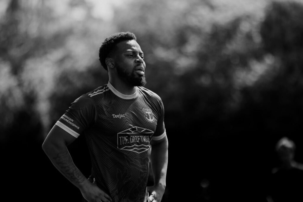 A football player stands with a focused expression, wearing a dark sports jersey. The background is blurred with natural scenery.