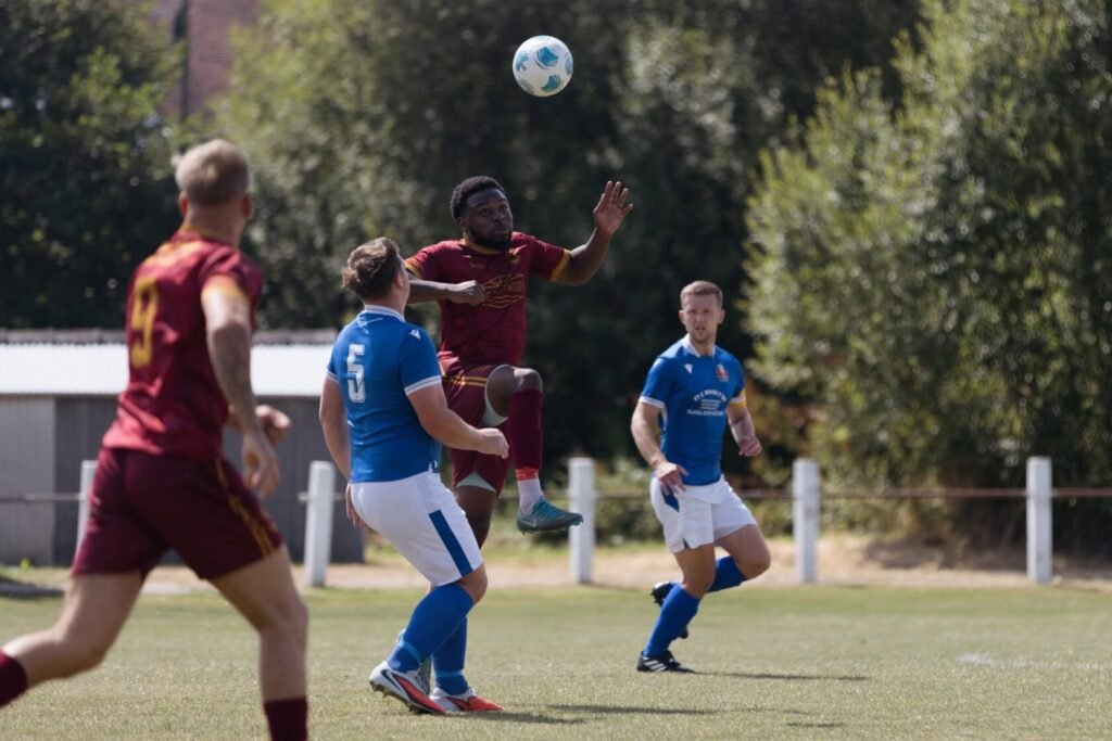 A player in red jumps to control a football in mid-air while two players in blue defend during a sunny football match.