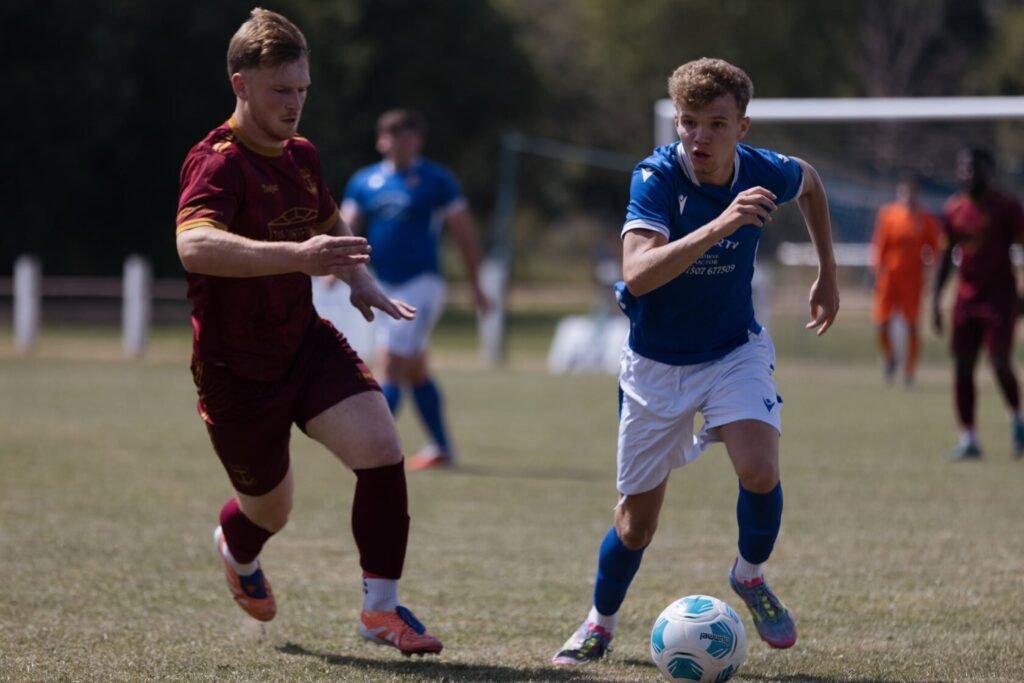 Two male soccer players competing for the ball on a grassy field; one in a maroon kit and the other in a blue kit.