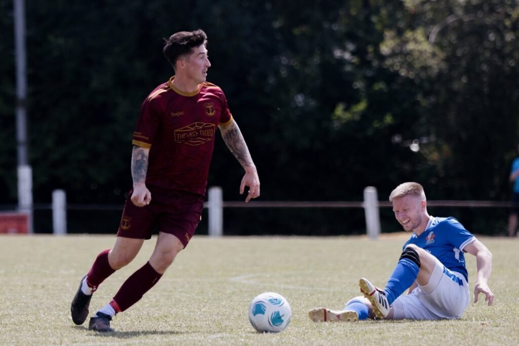 A football player in a maroon kit runs past another player in a blue kit, who is sitting on the ground with a smile. A football lies between them on the grassy field.