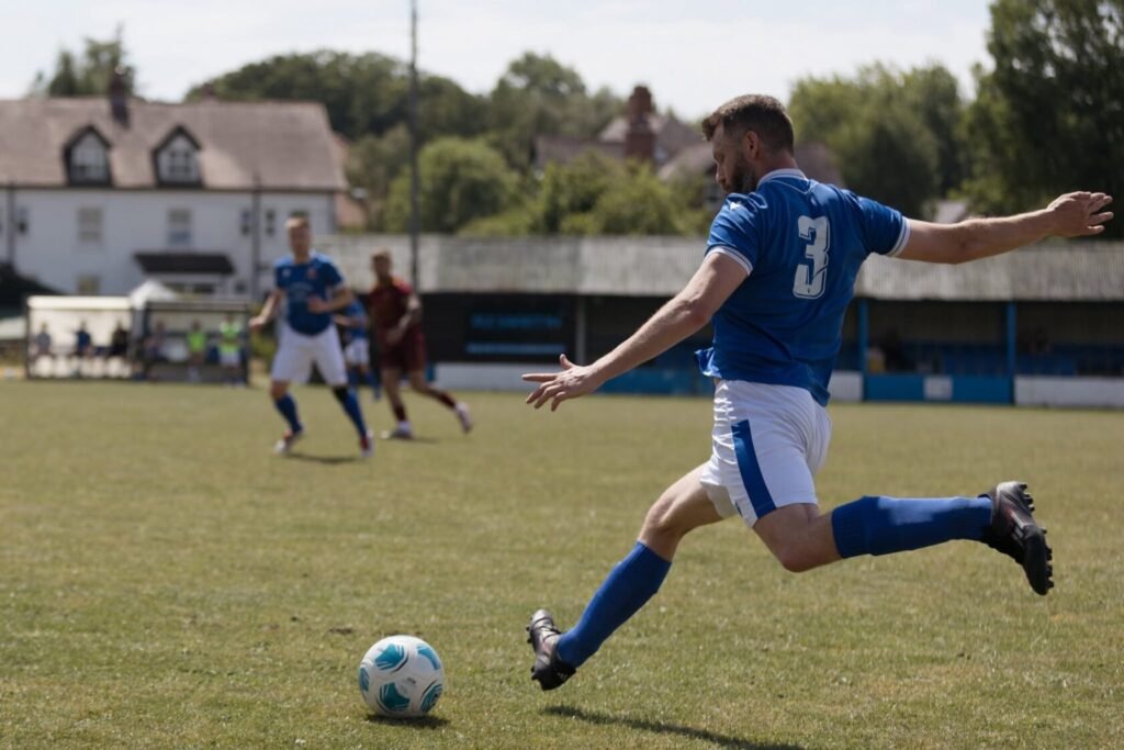 A male football player in a blue kit kicks a ball on a grassy pitch, with teammates and opponents in the background.