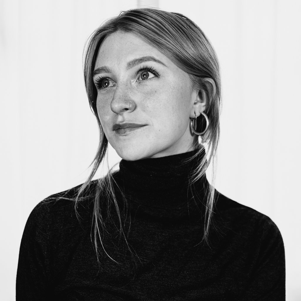 Close-up portrait of a woman with fair skin and long, light hair wearing a black turtleneck and hoop earrings, looking thoughtfully off-camera.