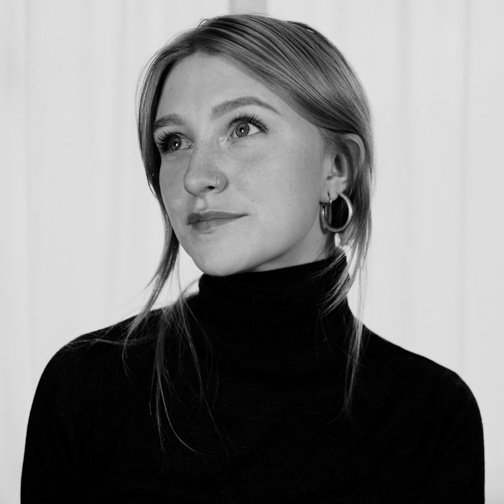 A young woman with blond hair and hoop earrings, wearing a black turtleneck, gazes thoughtfully upwards in a black and white setting.