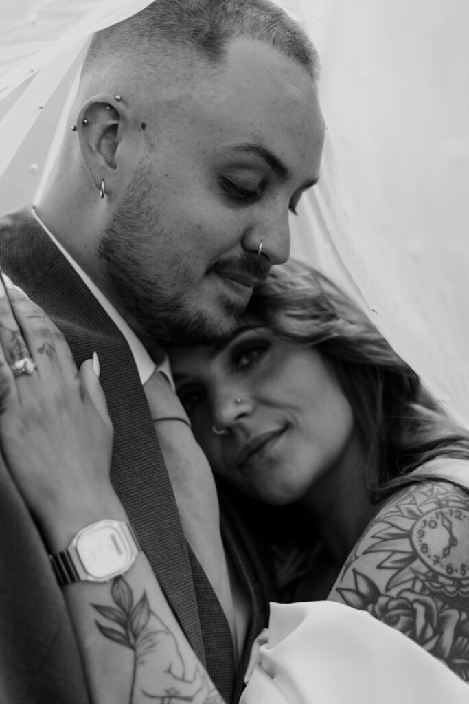 Close-up of a couple embracing under a veil, featuring intricate tattoos and wedding rings, captured in black and white.