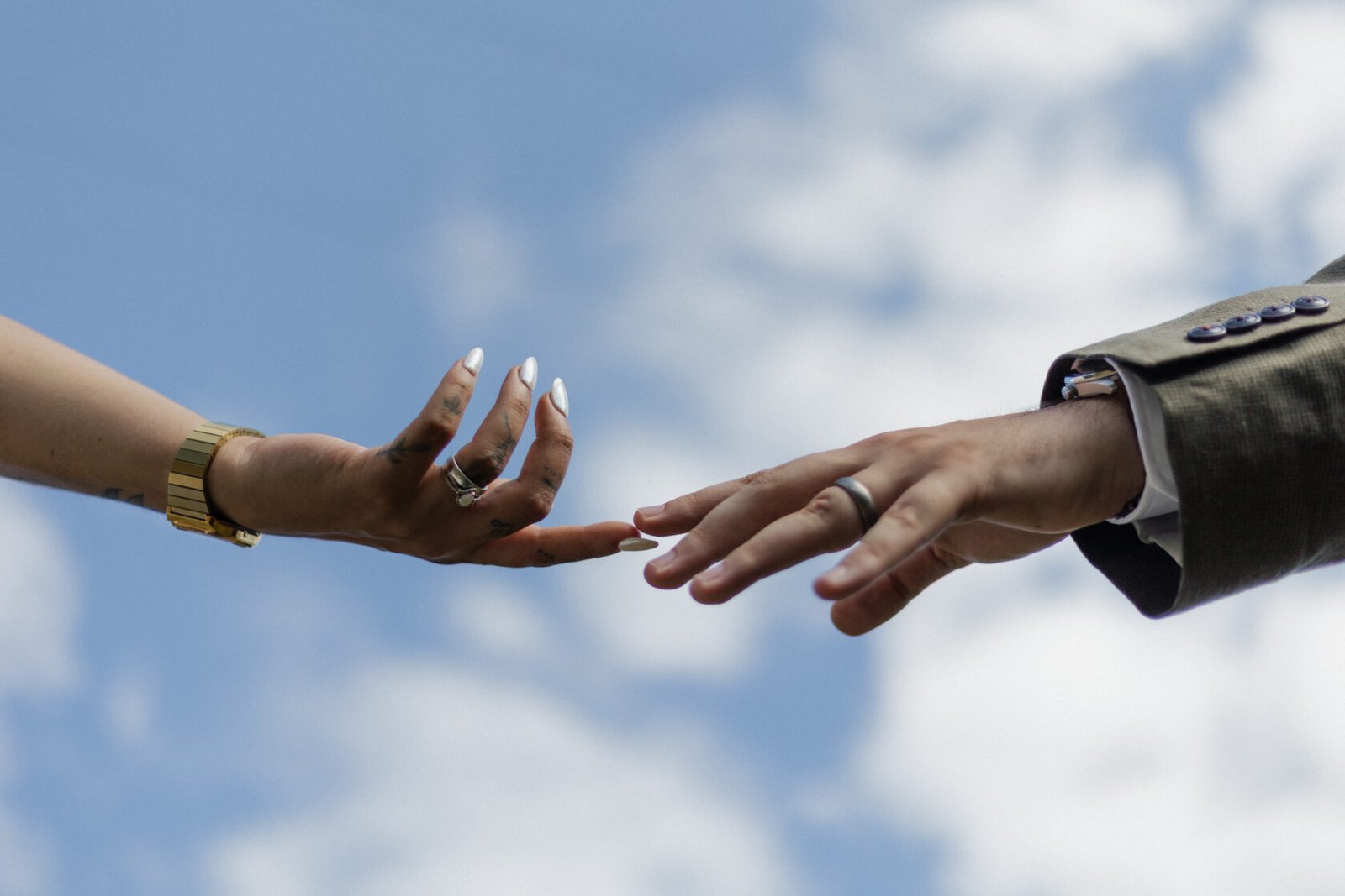 Two hands reaching out to each other with a bright blue sky and clouds in the background. One hand has tattoos and rings, while the other is dressed formally with a suit.