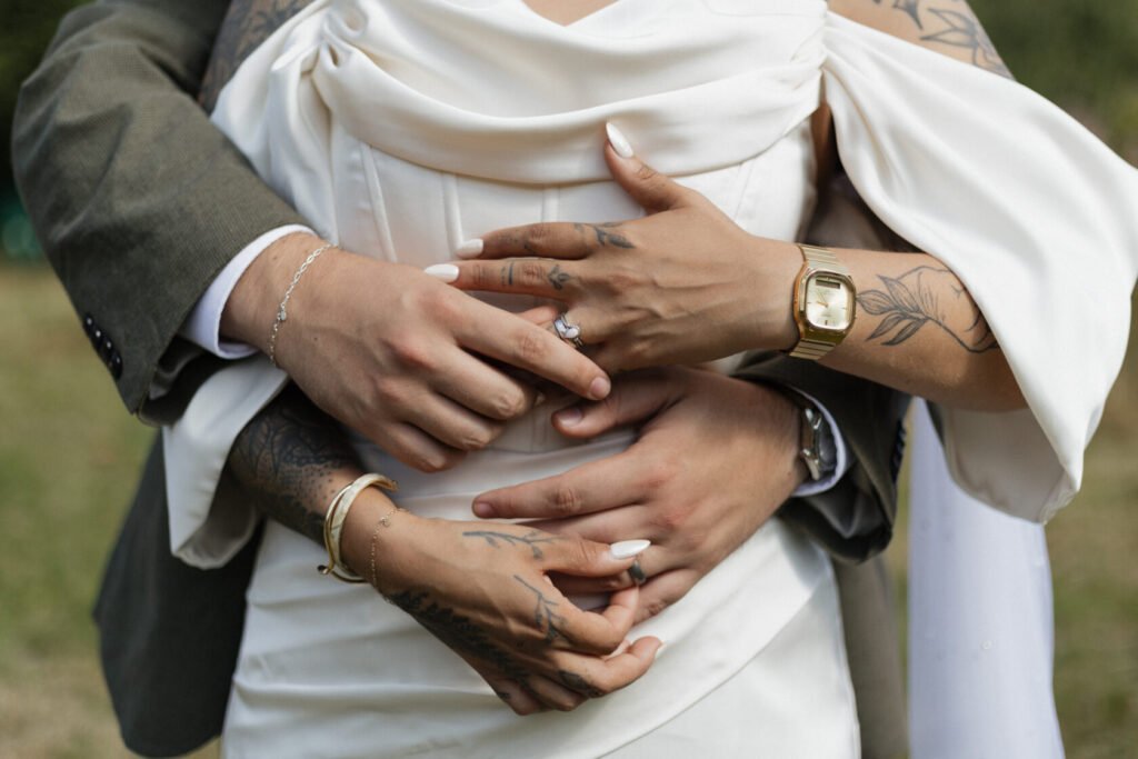 Close-up of a couple's intertwined hands, showcasing elegant wedding rings and intricate tattoos, dressed in a formal wedding attire.