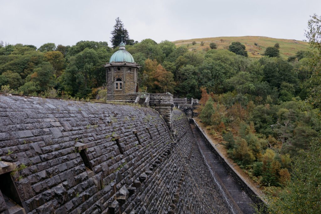 A stone dam with a round tower and a green dome, set against a backdrop of trees and hills under a cloudy sky.