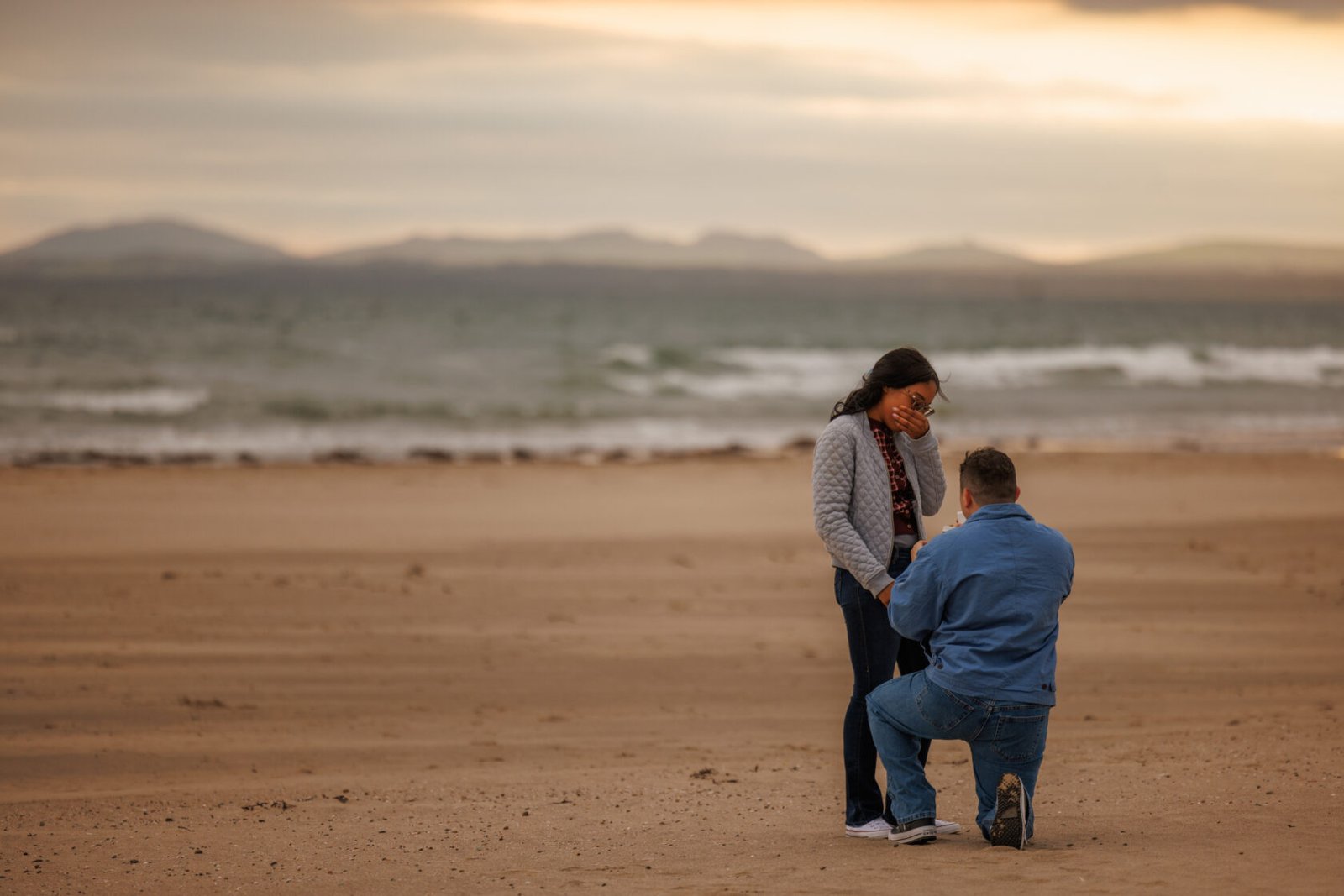 A man kneels on a sandy beach, holding a ring box, while a woman smiles and covers her mouth in surprise, with ocean waves and mountains in the background.
