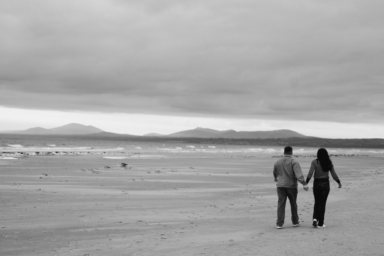 A couple holding hands while walking on a sandy beach, with mountains in the background and a cloudy sky.