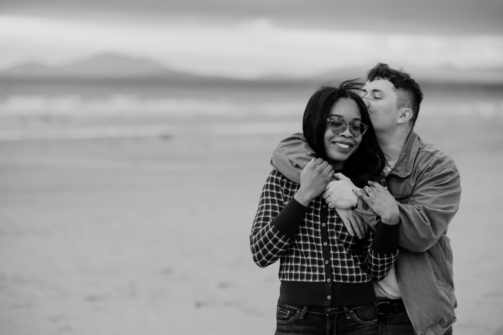 A couple sharing an affectionate embrace on a beach, with the woman smiling and the man kissing her neck. They are dressed casually, with a scenic ocean background.
