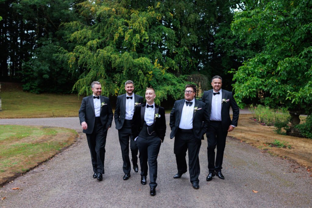 Five groomsmen walking together on a gravel path, dressed in tuxedos with black bow ties and floral boutonnieres, surrounded by greenery.