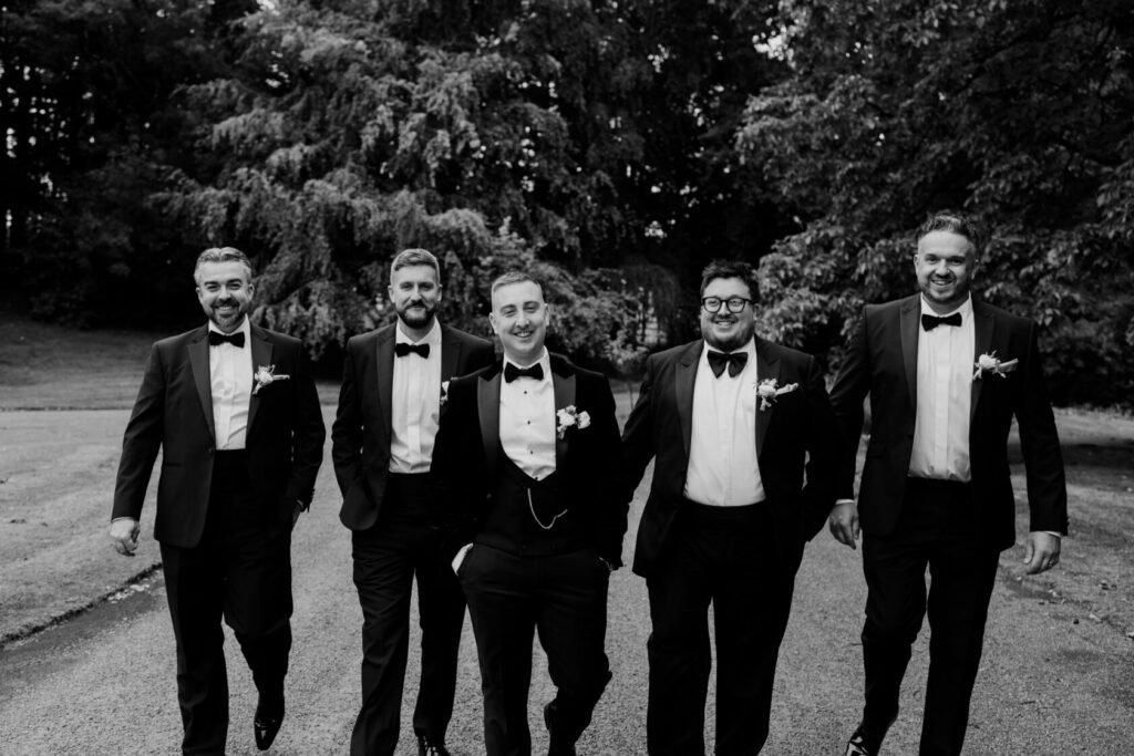 A group of five men dressed in tuxedos walking together, smiling, surrounded by greenery.
