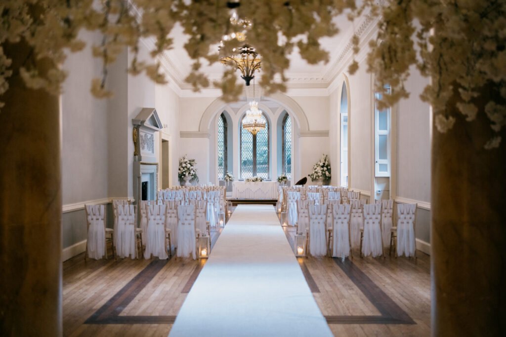 Interior of a wedding venue with rows of white chairs, a central aisle, and floral decorations.