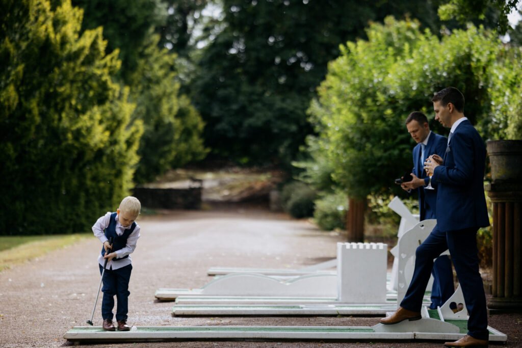 A young boy in a formal outfit is playing mini golf while two men in suits stand nearby, one holding a drink. Green trees frame the scene in a well-maintained outdoor area.