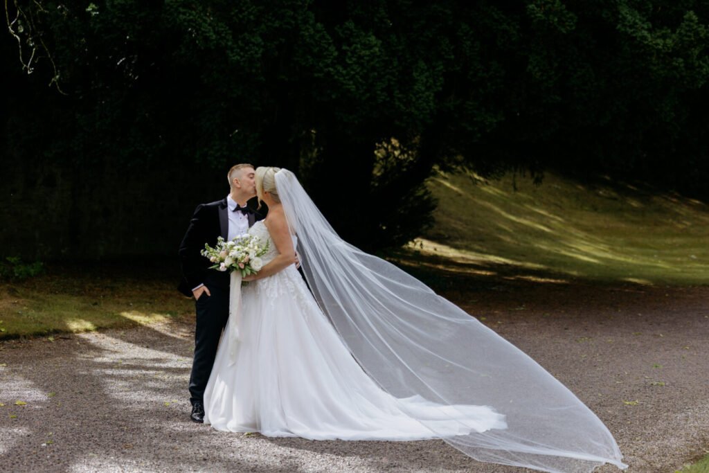 Bride and groom sharing a kiss outdoors, with the bride wearing a flowing white wedding gown and a long veil, while holding a bouquet.