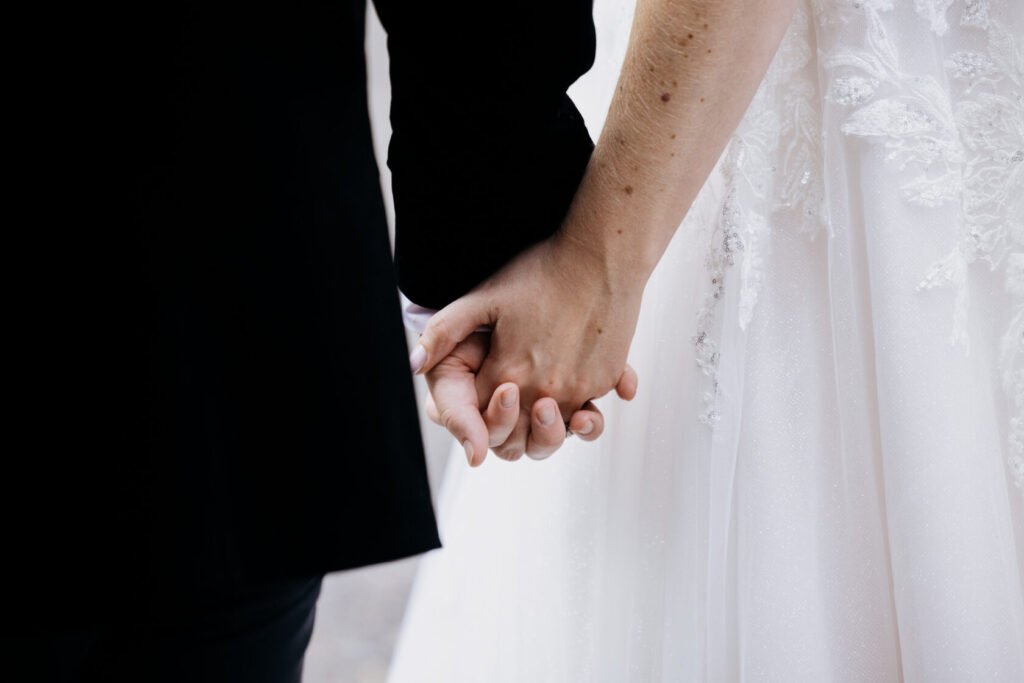 Close-up of a couple holding hands, one wearing a formal black outfit and the other in a white wedding dress with intricate detailing.