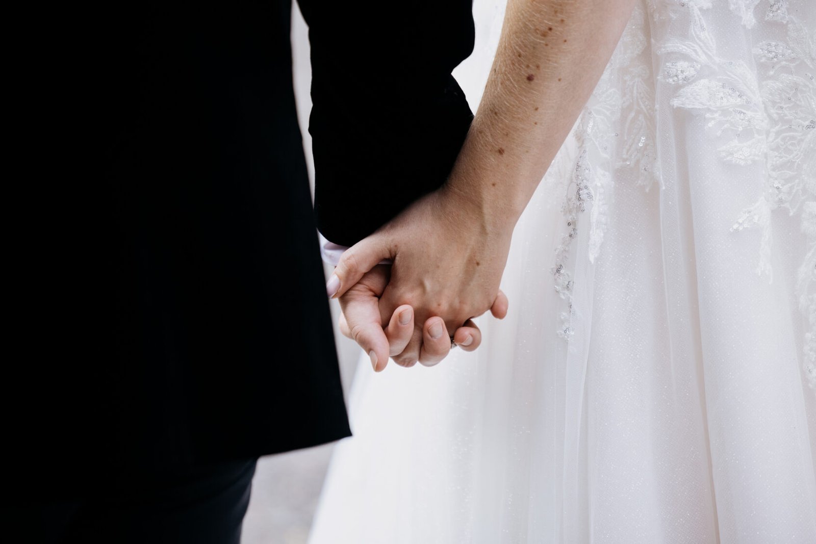 Close-up of a couple holding hands, one wearing a formal black outfit and the other in a white wedding dress with intricate detailing.