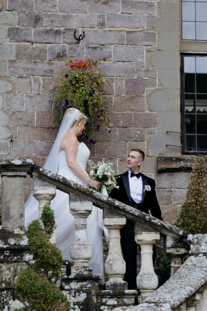 A bride in a white wedding gown and a groom in a black tuxedo share a moment on a stone staircase, with flowers in the background.