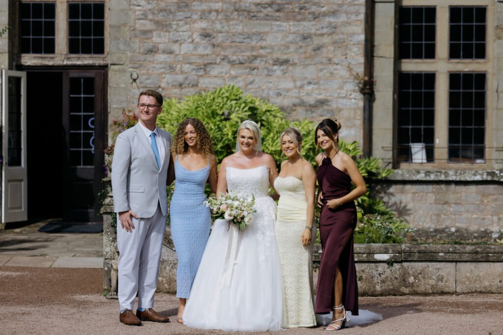 A smiling bride in a white gown stands with four guests, including two women in colourful dresses and a man in a light suit, against a stone building.