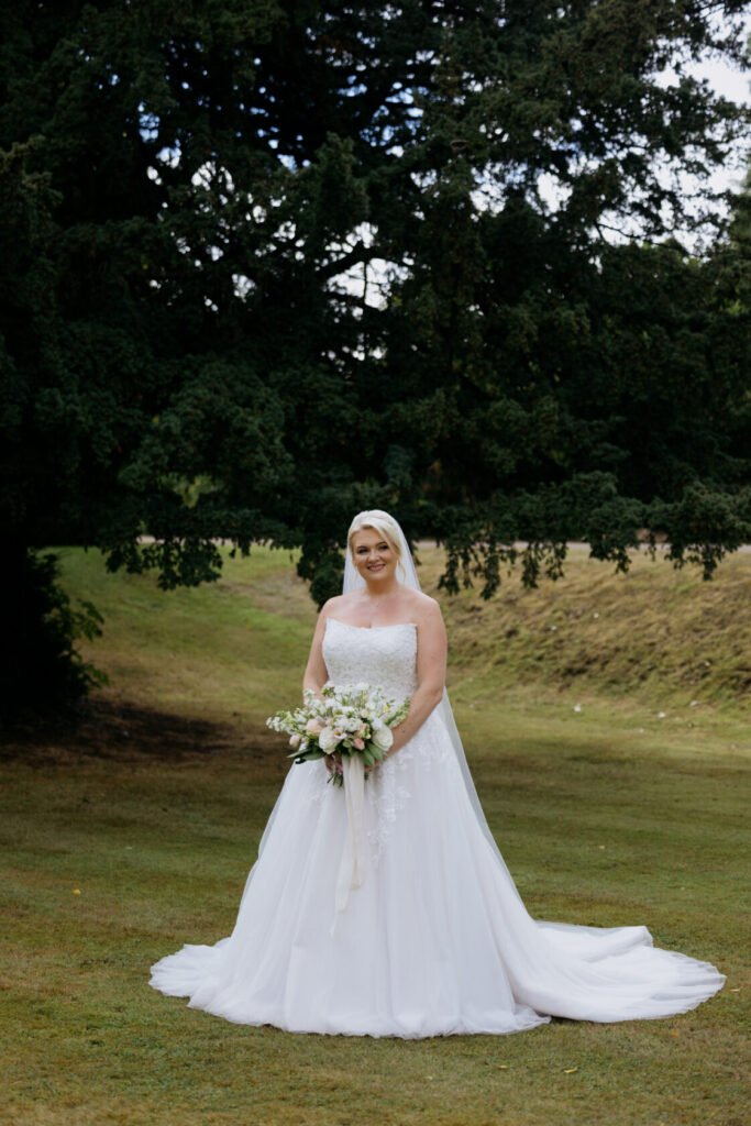 A smiling bride wearing a white wedding dress and veil, holding a bouquet of flowers, standing on grass with trees in the background.