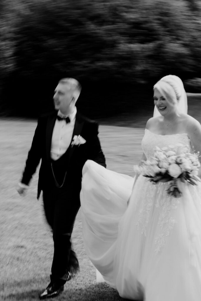 A blurred black and white photograph of a groom in a tuxedo holding the hand of a bride in a flowing wedding dress, both smiling as they walk together outdoors.