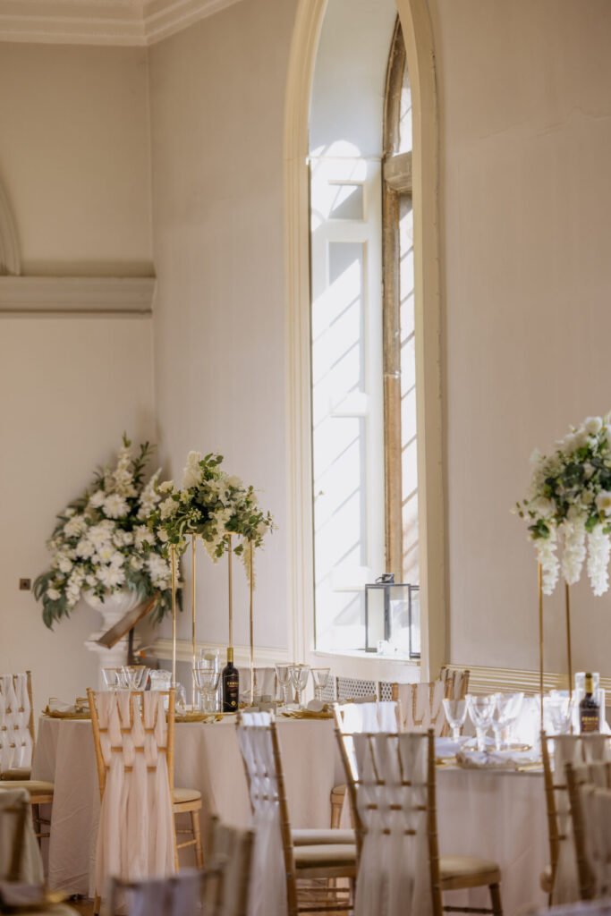 A beautifully arranged wedding reception with tables set for dining, featuring floral centrepieces and glassware, illuminated by natural light from large windows.