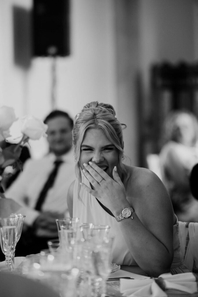 A woman laughing with her hand over her mouth at a dining table, surrounded by glassware and flowers, with a man in the background smiling.